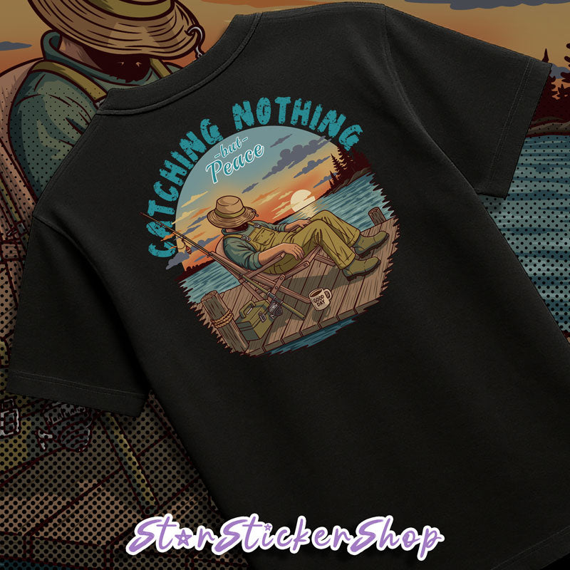 A black short-sleeve t-shirt laid flat, showing a large circular graphic on the back. The graphic features a man relaxing on a wooden dock by a lake at sunset, with the text 'CATCHING NOTHING But Peace' arched above him.