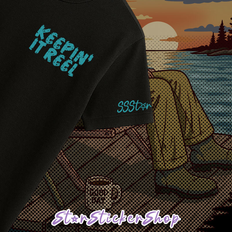 A close-up of a black t-shirt featuring a stylized graphic print. The graphic shows a retro sunset over water with a person sitting on a dock, alongside turquoise text reading 'KEEPIN' IT REAL' on the chest and 'SSStax' on the sleeve.