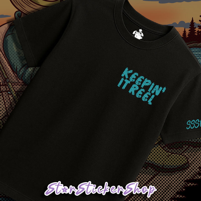 A black short-sleeve t-shirt with a distressed turquoise graphic reading 'KEEPIN' IT REEL' on the chest, displayed on a patterned surface with a scenic background.
